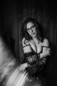 Nő fekete-fehér boudoir fotón, csipkés köntösben és szemüvegben pózolva – önbizalomnövelő fehérneműs fotózás természetes hangulatban.