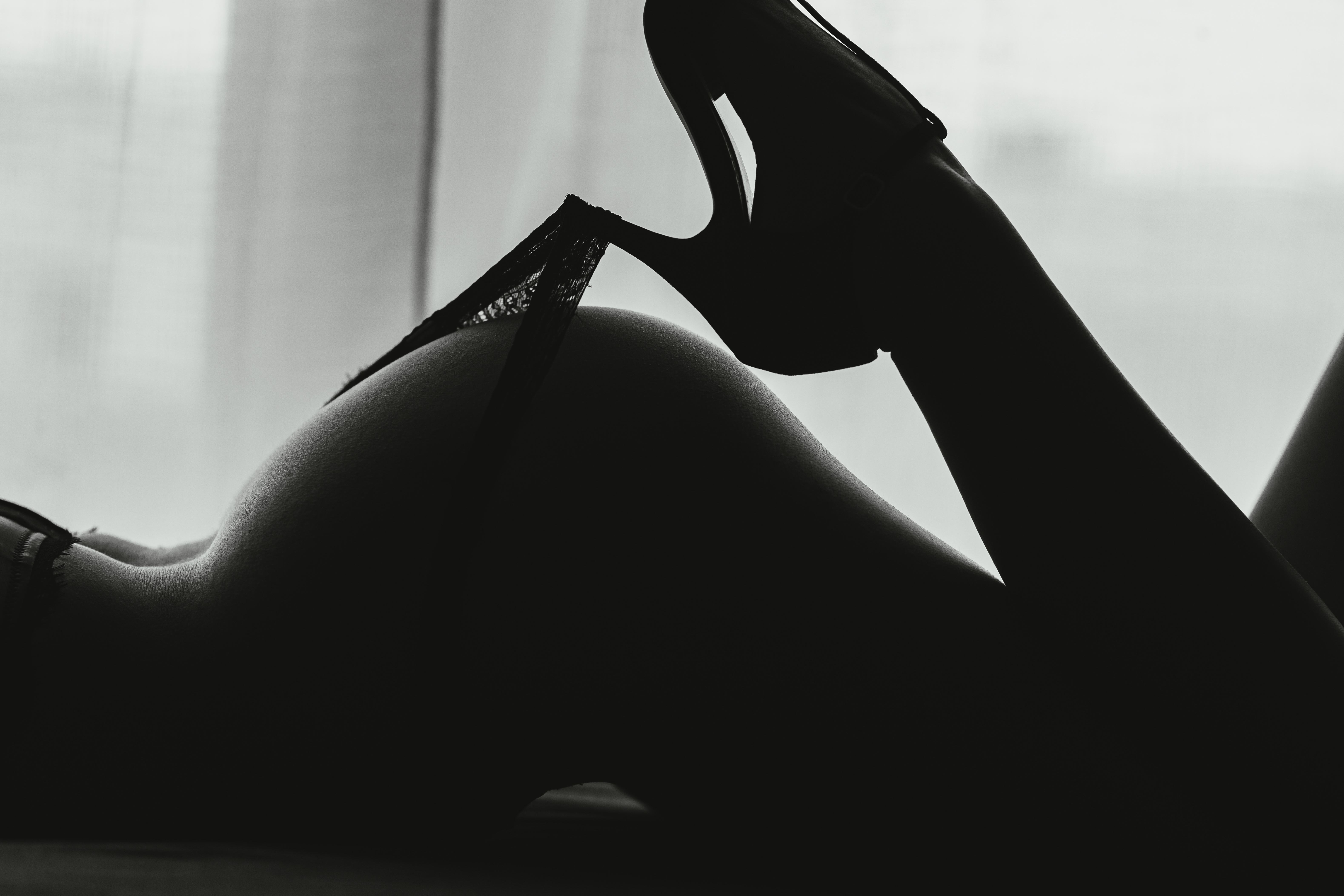 Erotikus fekete-fehér sziluettfotó női alakról, csipkebugyit lehúzva magas sarkú cipővel az ablak fényében.